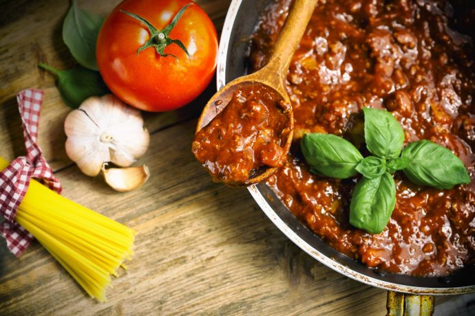 Bowl of bolognese sauce, with tomato, garlic and dried pasta on table.